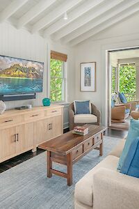 The image features a bright, cozy living room with a TV, wooden furniture, and large windows showing lush greenery outside.