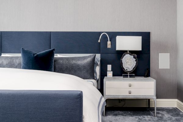 A modern bedroom features a blue headboard, white bedding with blue pillows, and a nightstand with a lamp, clock, and artwork on a blue carpeted floor.