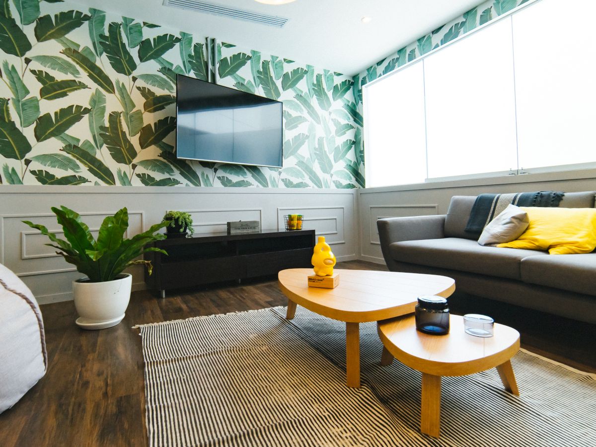 A modern living room with a tropical leaf wallpaper, gray sofa, yellow pillows, wooden coffee table, and a potted plant near the TV stand.