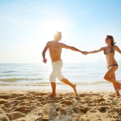 A man and woman holding hands are running on a sandy beach near the ocean under a bright sky.