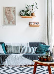 A cozy room with a sofa, pillows, plants, a coffee table, wall art, and ample natural light from large windows. Shelves with decor adorn the wall.