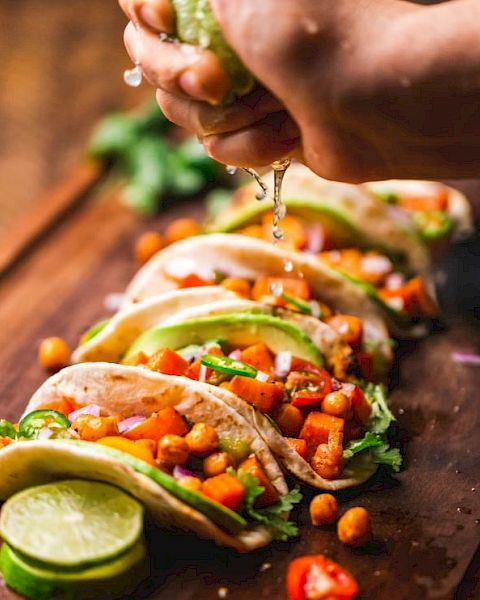 The image shows a hand squeezing a lime over a row of colorful tacos filled with vegetables and chickpeas on a wooden surface.