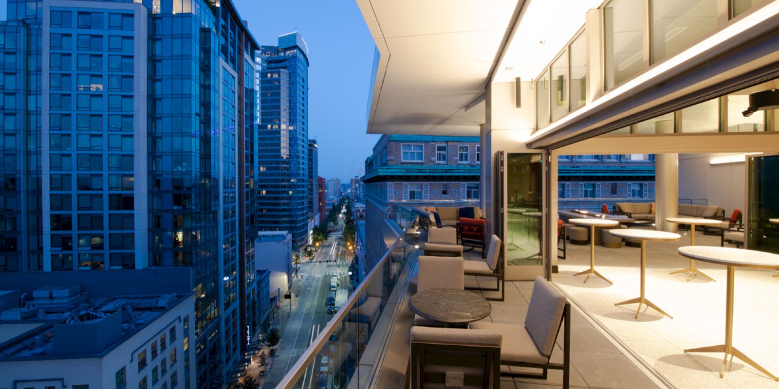 This image shows a modern balcony with tables and chairs overlooking a cityscape at dusk, featuring tall buildings and illuminated streets.