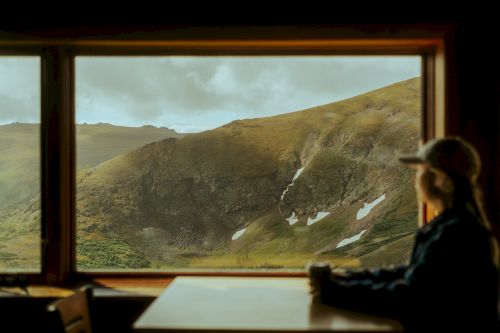 A person sits at a table, gazing out a large window at a hilly landscape with patches of snow, creating a peaceful and scenic view.