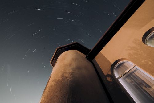 The image shows a night sky with star trails above a building tower and an adjacent window, indicating a long-exposure photograph.