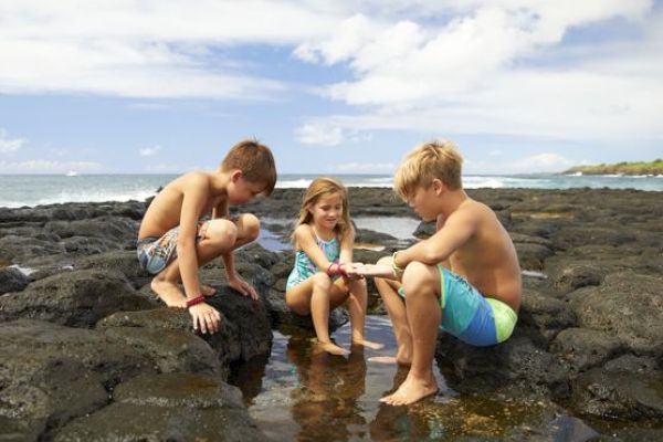 Three children in swimsuits are exploring a tidal pool on a rocky shore under a partly cloudy sky.