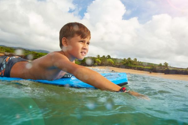 A young boy is lying on a blue surfboard in the ocean, looking towards the shore under a partly cloudy sky with sunlight shining.