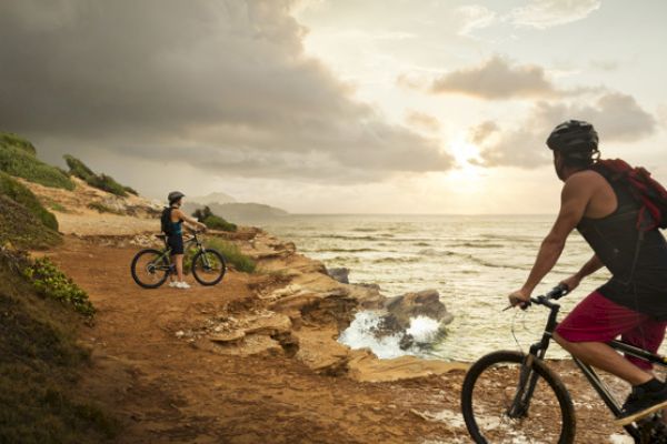 Two people are riding bicycles on a rocky coastal path, with a scenic view of the ocean and an overcast sky in the background.