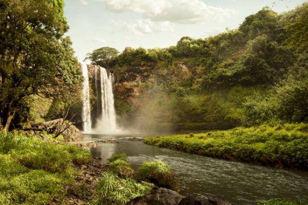 A picturesque waterfall cascades into a serene river, surrounded by lush greenery and trees under a partly cloudy sky.