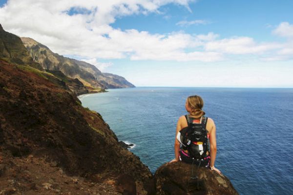 A person with a backpack sits on a rocky cliff overlooking the ocean, with a dramatic coastline and partly cloudy sky in the background.