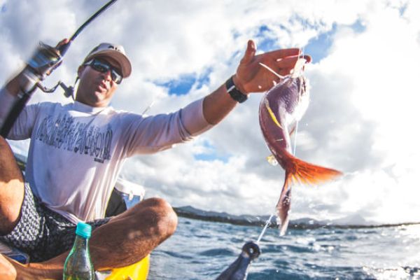 A person is fishing from a yellow kayak, holding a fishing rod and a colorful fish they've just caught, with the ocean and a cloudy sky in the background.