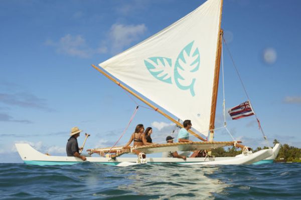 A group of people is sailing on a traditional outrigger canoe with a large sail, on open waters, under a clear sky with land visible in the background.