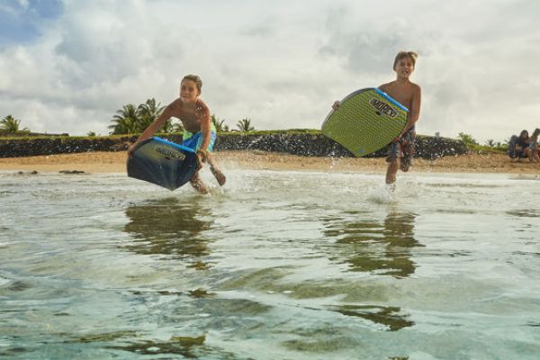 Two children are running into the ocean with bodyboards on a sunny day at the beach, with some people and palm trees in the background.