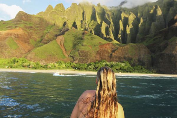 A person with long hair, wearing a swimsuit, is seen from behind looking at a scenic coastal mountain landscape.