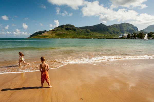 Two children are playing on a sandy beach with clear water and distant green hills under a bright blue sky with scattered clouds.