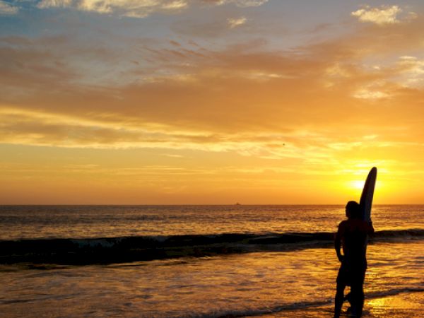 A person standing on the beach holding a surfboard is silhouetted against a vibrant sunset sky with clouds reflecting on the calm ocean water, ending the sentence.