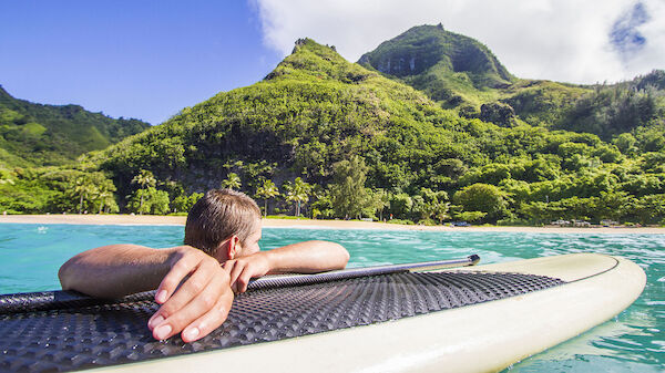 A person is lying on a surfboard in clear water, looking towards lush green mountains and a beach under a blue sky with some clouds.