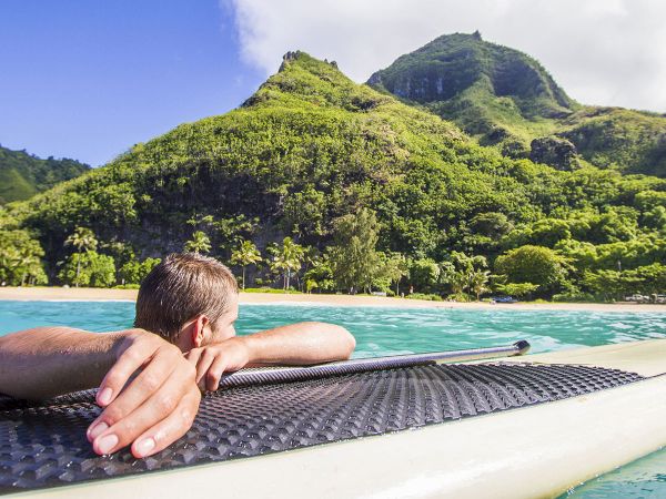 A person is lying on a surfboard in clear water, looking towards lush green mountains and a beach under a blue sky with some clouds.