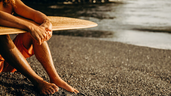 A person with tanned skin sits on a sandy beach, holding onto a surfboard, and looking out towards the ocean in the background.