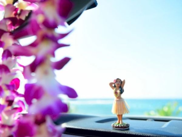 A hula dancer figurine on a car dashboard, with purple flowers and a blue ocean in the background.