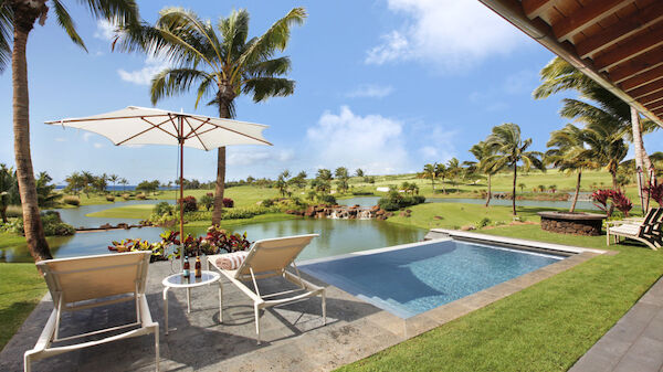 A serene outdoor setting with a pool, sun loungers, an umbrella, palm trees, a pond, and expansive green fields under a blue sky with some clouds.