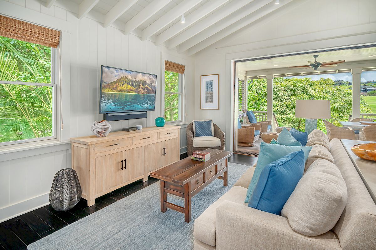 The image shows a modern living room with comfortable furniture, a TV, and large windows overlooking greenery. It has a light, airy feel.