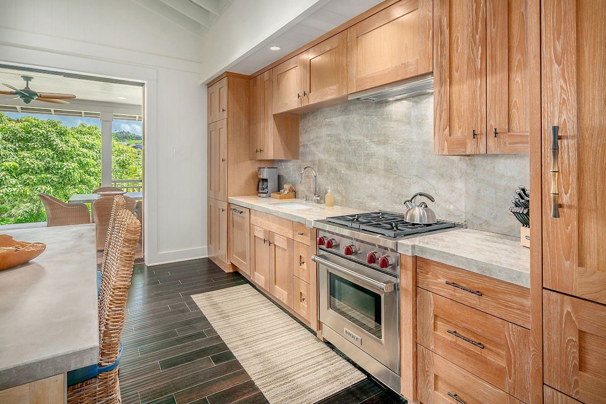 The image shows a modern kitchen with wooden cabinets, a gas stove, and a calming view outside through large windows.