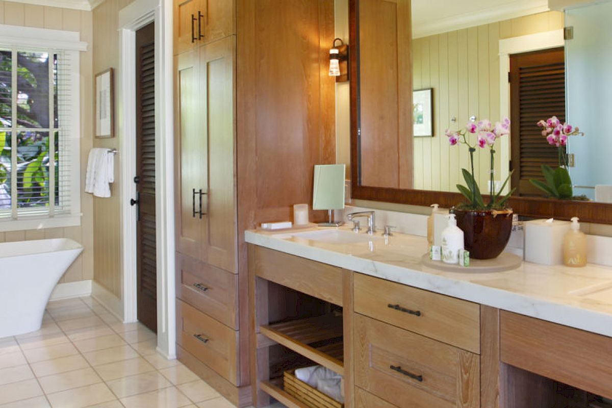 A spacious bathroom featuring a freestanding bathtub by the window, a double vanity with a large mirror, wooden cabinets, and potted flowers on the counter.