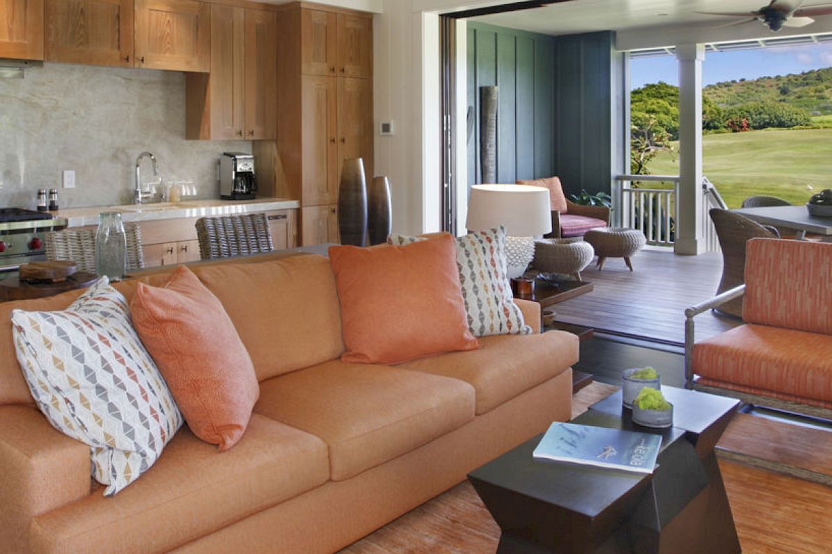 A cozy living area with a kitchen, orange couch, and open patio doors leading to an outdoor seating area with scenic views of a green landscape.