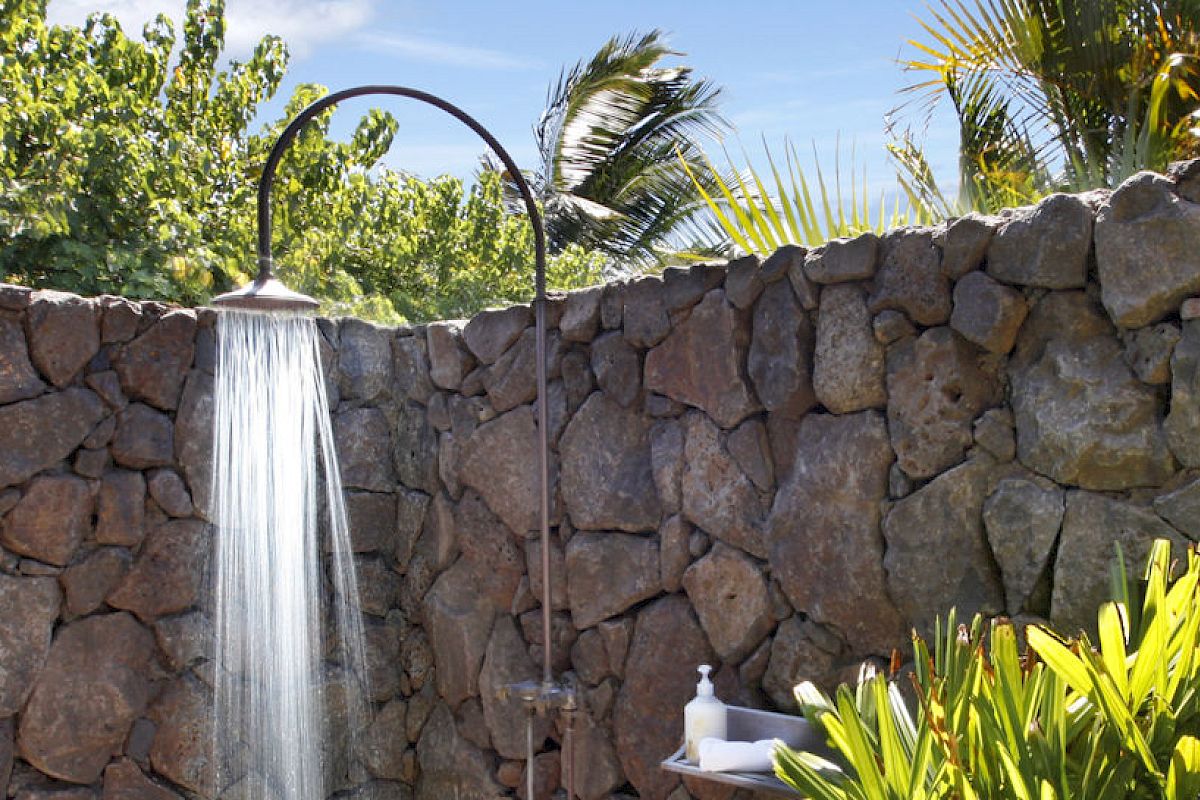An outdoor shower with water flowing, set against a stone wall, lush greenery, and clear sky in the background.