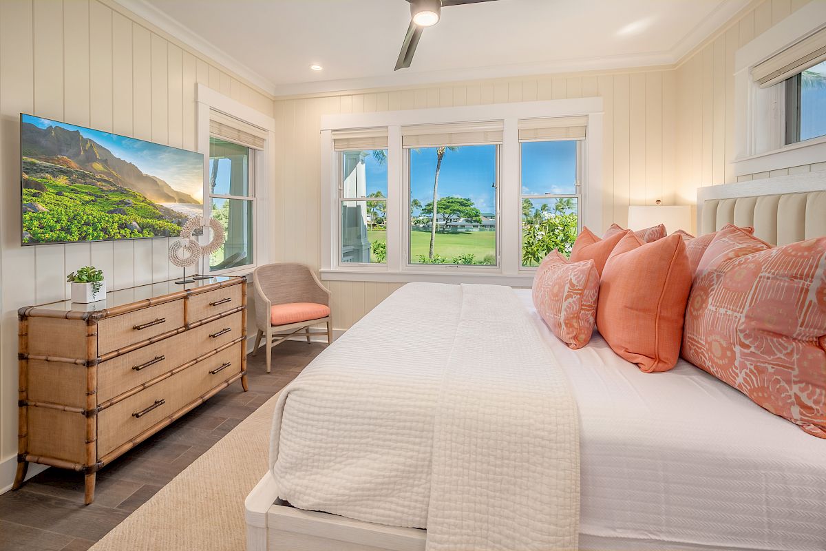 A cozy bedroom features a large bed with pink pillows, a wooden dresser with a TV, a chair, and windows revealing an outdoor view.