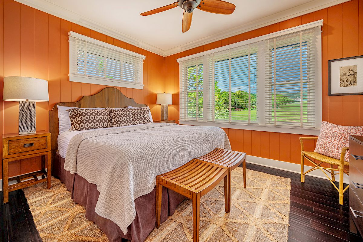 The image shows a cozy bedroom with orange walls, a queen bed, wooden furniture, two lamps, and large windows with a view.