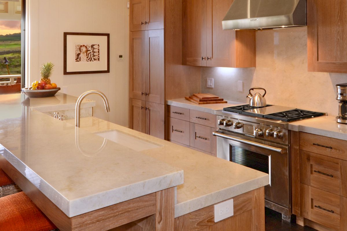 A modern kitchen with wooden cabinets, a marble countertop, stainless steel appliances, and bar seating, opens to an outdoor patio with a scenic view.
