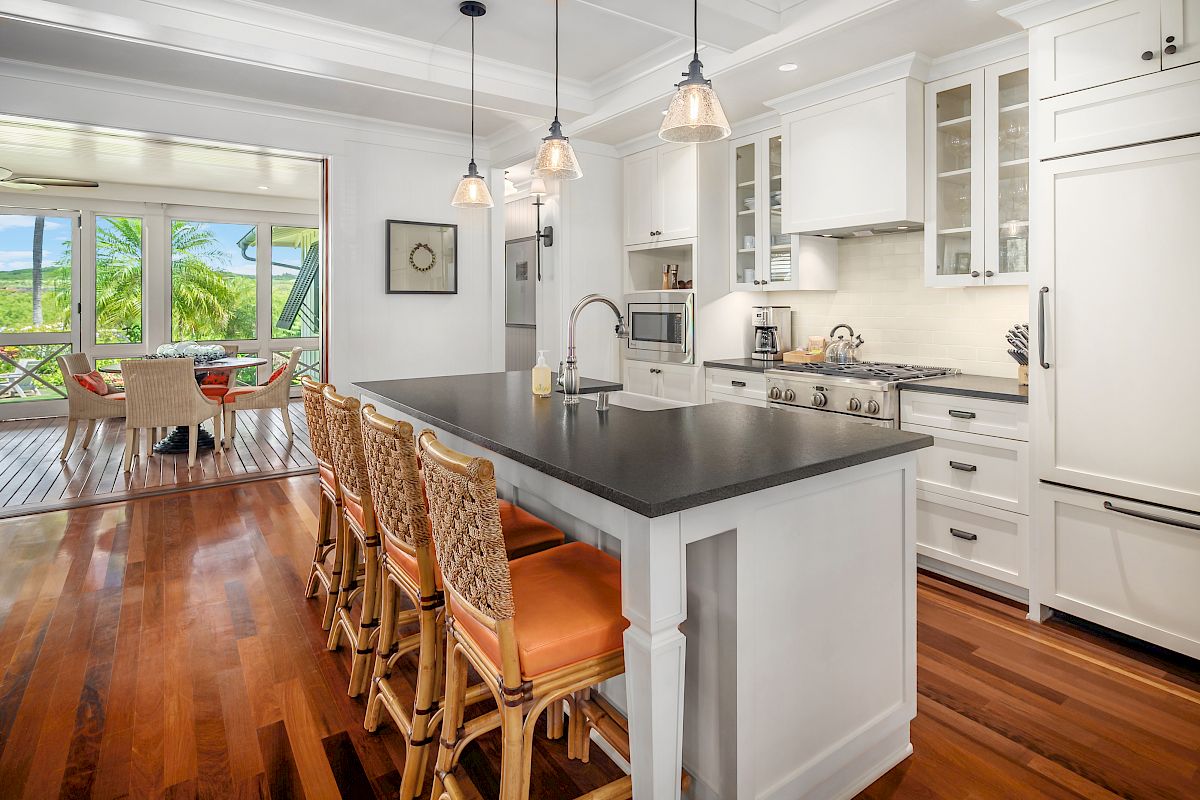 A modern kitchen with a large island, bar stools, white cabinets, hanging lights, and a view of greenery through sliding doors.