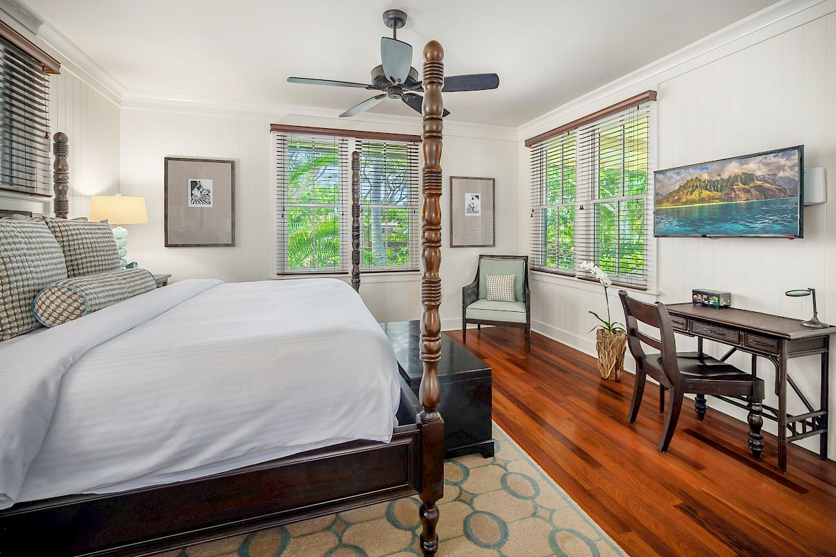 The image shows a cozy bedroom featuring a large bed, wooden flooring, a work desk, and windows with greenery outside.
