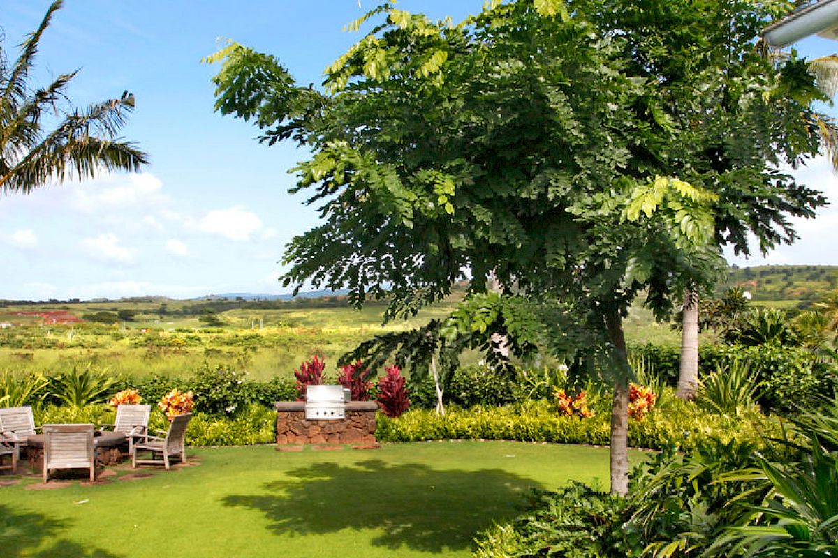 A lush garden with a seating area, palm trees, and vibrant flowers, overlooking a scenic landscape under a clear, blue sky.