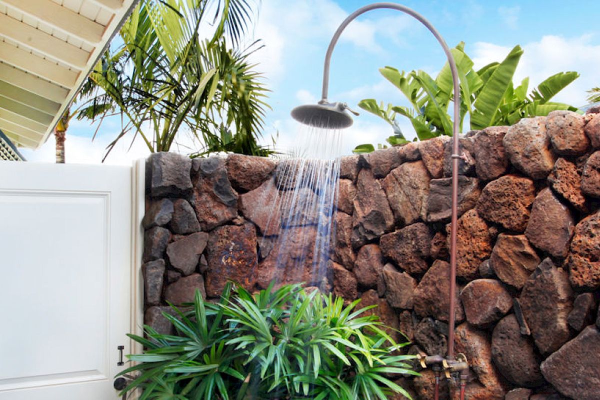The image shows an outdoor shower with stone walls, surrounded by lush greenery and a tropical ambiance, including palm trees and clear skies.