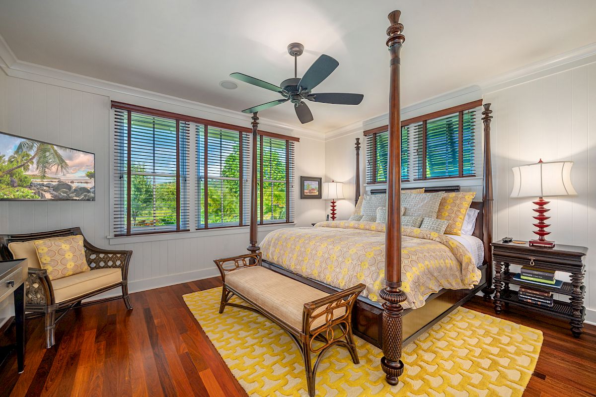 A bedroom with a four-poster bed, yellow bedding, ceiling fan, TV, chairs, a bench, nightstands, lamps, and large windows letting in natural light.