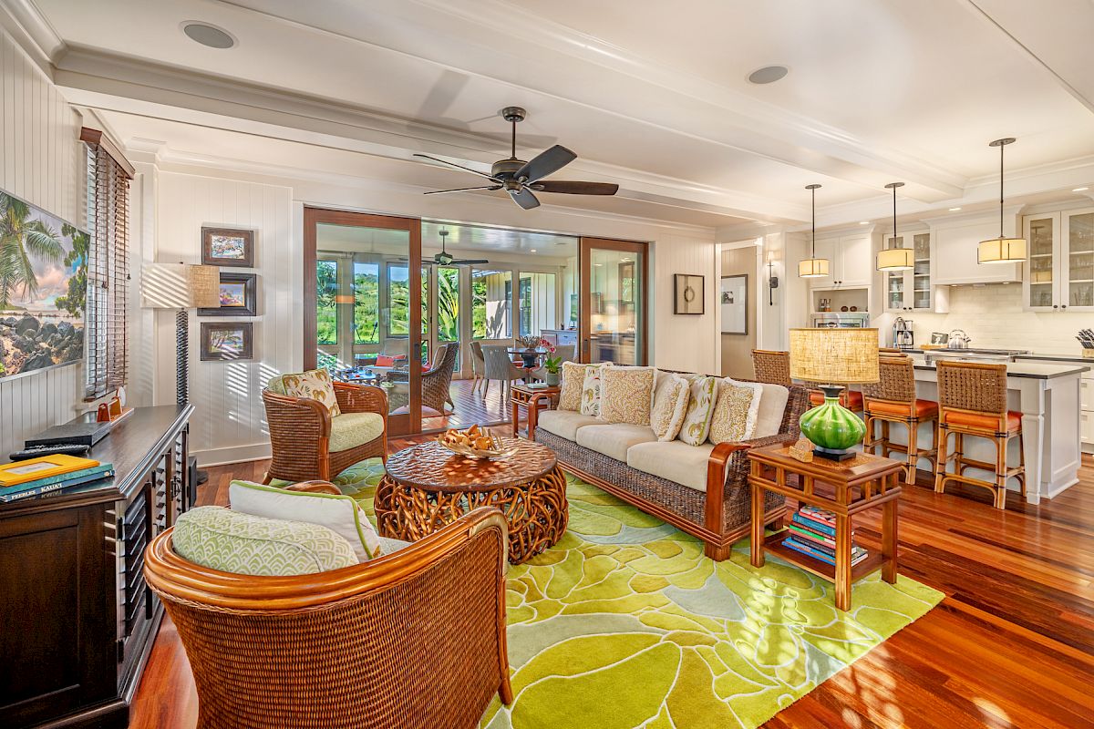 A cozy living room with wicker furniture, a green rug, and a ceiling fan adjacent to a bright kitchen; large sliding doors open to the patio.