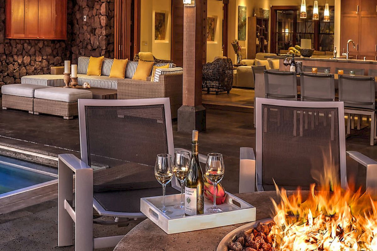 An outdoor patio with a fire pit, pool, wine glasses, lounge chairs, and an open view into a cozy indoor living and kitchen space.