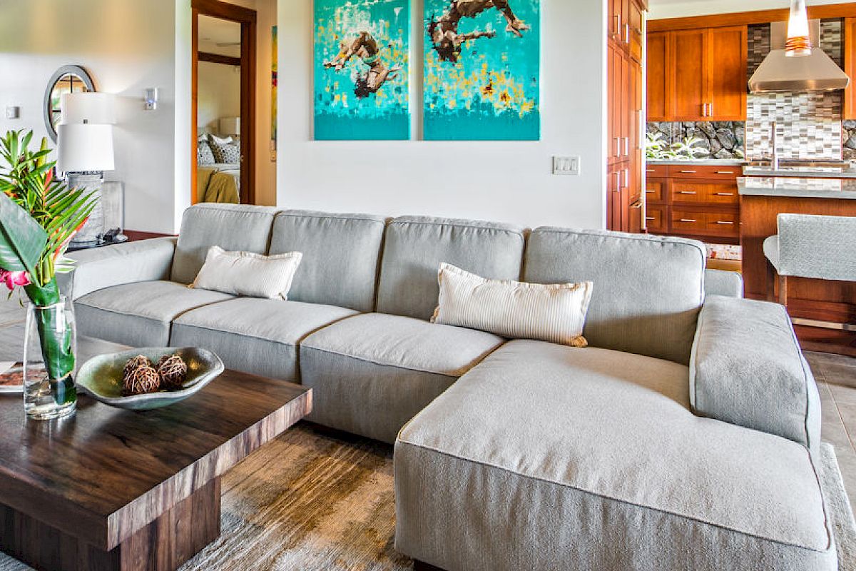 A modern living room with a gray sectional couch, a wooden coffee table, and a kitchen with wooden cabinets and barstools in the background.