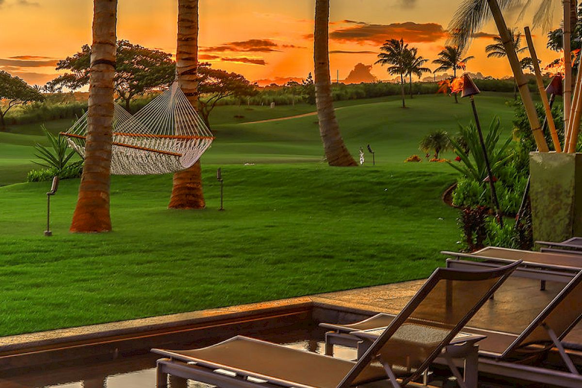 The image shows a tranquil outdoor scene with lounge chairs, a hammock between palm trees, and a lush green lawn during a beautiful sunset.