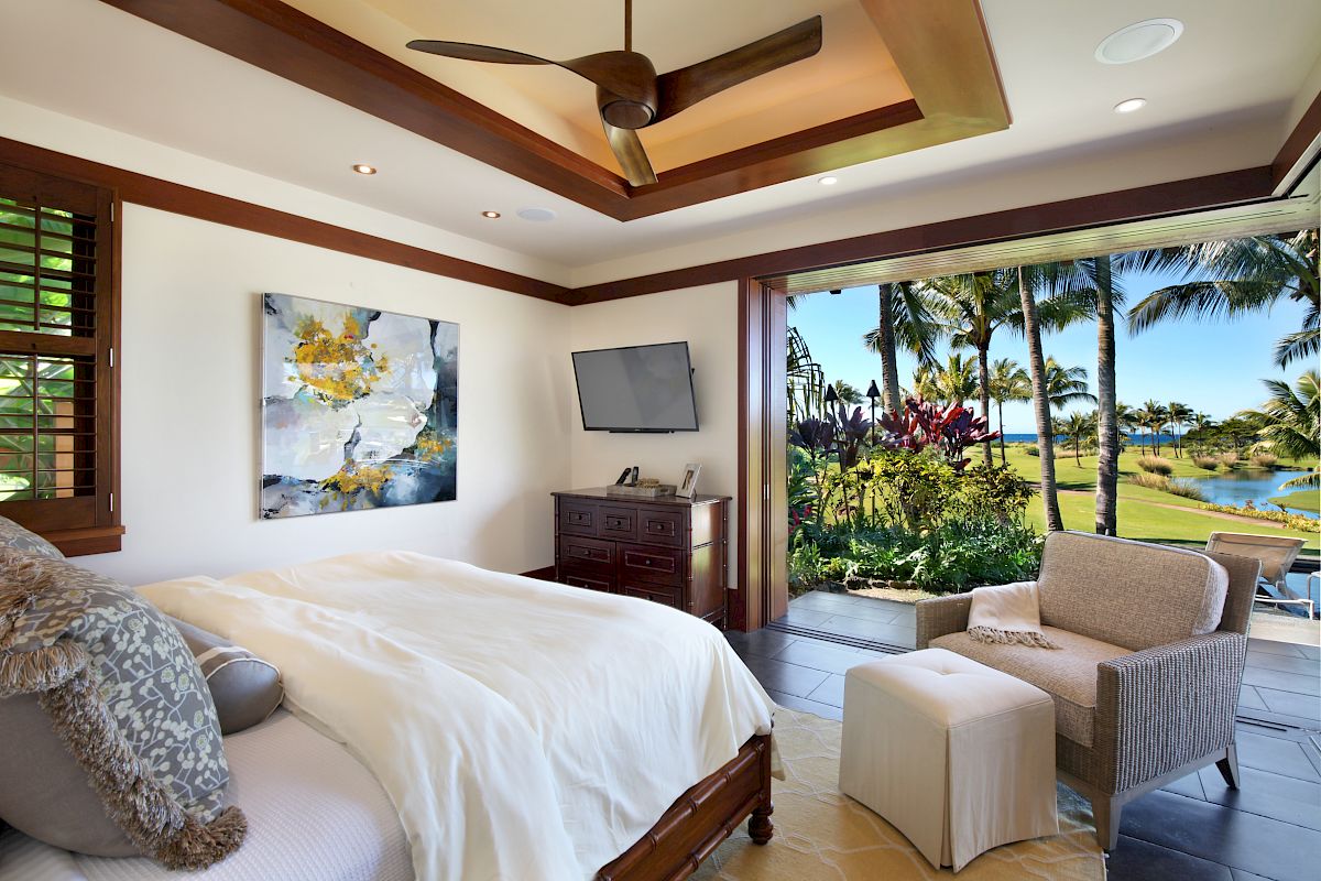 A bedroom with a ceiling fan, painting, TV, armchair, and ottoman leads to an open patio with a view of palm trees and a pond.