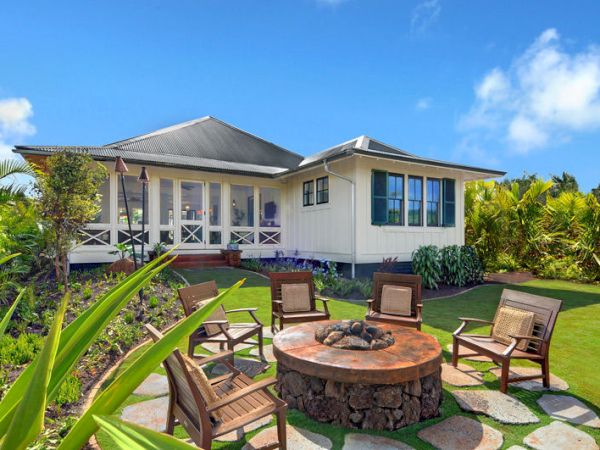 A cozy outdoor setup with a central fire pit, surrounded by chairs, located in a lush garden area in front of a quaint, single-story house.