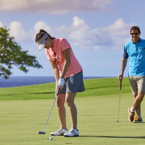 A woman is putting on a golf green while a man holds the flagstick nearby; both appear to be enjoying a sunny day on the golf course.