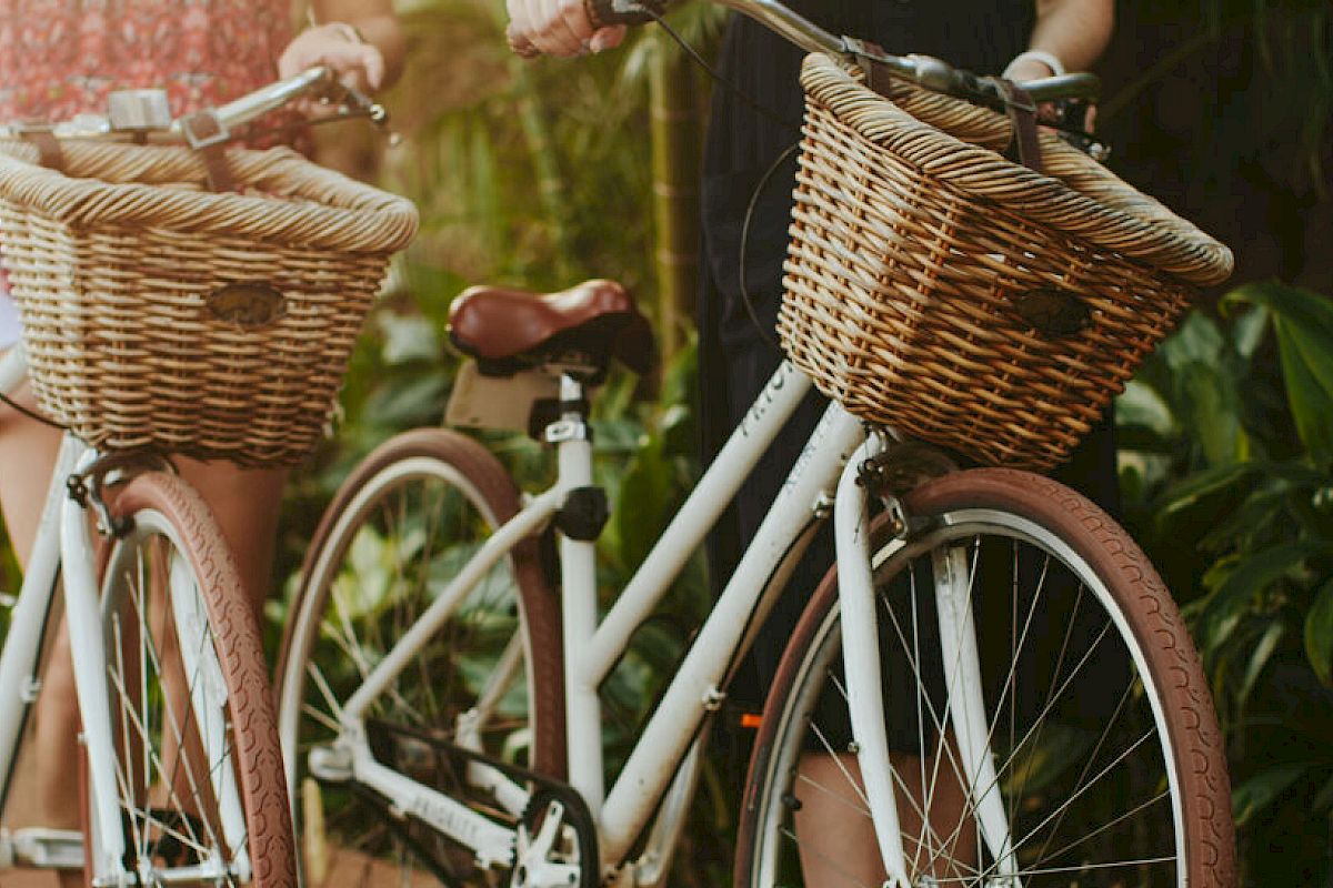 Two people are standing next to bicycles with wicker baskets in a lush, green outdoor setting.