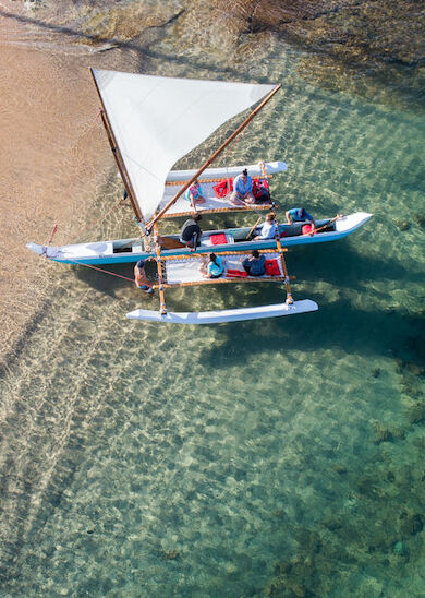 An aerial view of a sailboat with people on it, near a sandy shore in clear, shallow water on a sunny day.