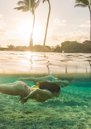 Two people snorkeling underwater in a clear tropical ocean, with the sun setting behind palm trees in the background.