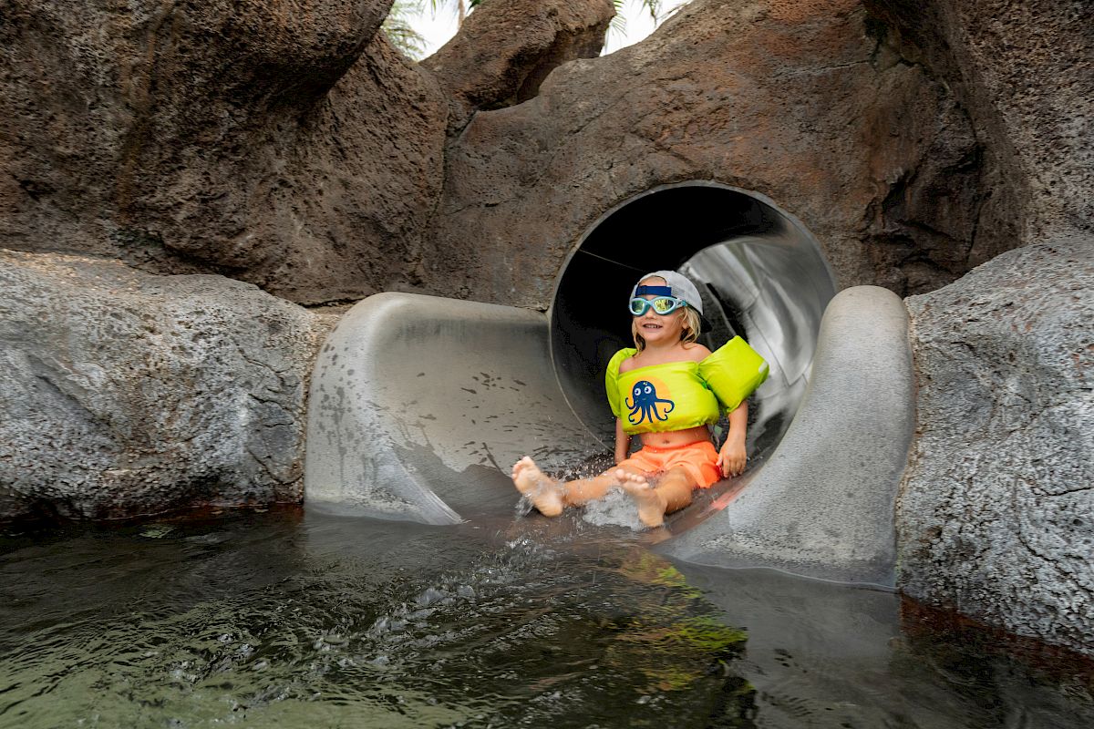 A child wearing swimming gear and a life vest is coming out of a water slide in a rocky setting, splashing into a pool.