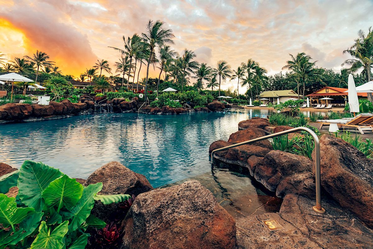 A serene pool surrounded by lush tropical plants, rock formations, and palm trees, with lounge chairs and umbrellas under a vibrant sunset sky.